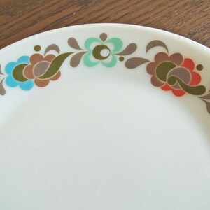 Peut inclure: Assiette en c&eacute;ramique blanche avec un motif floral. Les fleurs sont dans des tons de turquoise, marron, orange et vert olive. L'assiette a un bord incurv&eacute; et une surface lisse. Le motif floral est le long du bord de l'assiette.