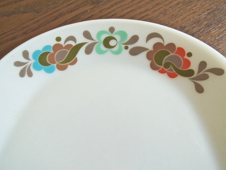 Peut inclure: Assiette en c&eacute;ramique blanche avec un motif floral. Les fleurs sont dans des tons de turquoise, marron, orange et vert olive. L'assiette a un bord incurv&eacute; et une surface lisse. Le motif floral est le long du bord de l'assiette.