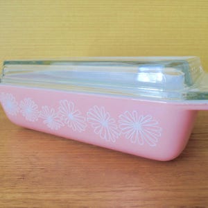 May include: A pink Pyrex baking dish with a clear glass lid. The dish features white daisy patterns on the sides. The dish is on a wooden surface. The dish is approximately 20 cm long.