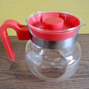 Peut inclure: Une carafe en verre transparent avec une poignée et un couvercle en plastique rouge. Une bande métallique argentée entoure le milieu de la carafe. La carafe est conçue pour servir des boissons.