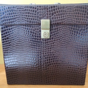 Könnte beinhalten: Eine dunkelbraune, rechteckige Box mit einer strukturierten Kunstleder-Krokodilmusterung. Sie hat einen Metallverschluss und ein Schlüsselloch. Die Box dient wahrscheinlich zur Aufbewahrung oder als Reisekoffer.