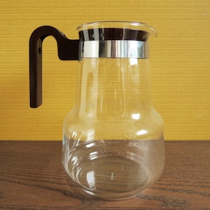 Peut inclure: Une carafe en verre transparent avec une poignée marron foncé et une bande argentée autour du haut. La carafe a une base arrondie et un dessus légèrement évasé. Le fond est de couleur jaune uni.