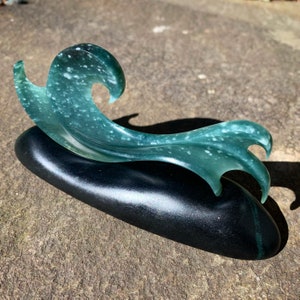 May include: A green wave sculpture with white flecks, carved from jade, resting on a black base.