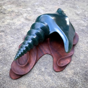 May include: A dark green, polished stone sculpture of a conch shell, mounted on a carved, reddish-brown wooden base. The shell's spiral and curved form are detailed. The sculpture is displayed on a textured, gray surface.