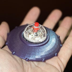 May include: A circular, metallic purple object shaped like a flying saucer with a shimmering finish and small protrusions along the edge. A clear, faceted dome sits in the center, topped with a small, solid red cylindrical button.