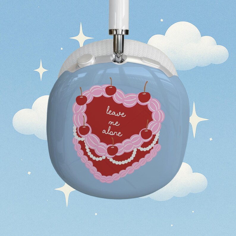 May include: Light blue over-ear headphones with a decorative heart-shaped cake design. The cake is red with pink frosting, cherries, and the text "leave me alone". The background is a blue sky with clouds and stars.