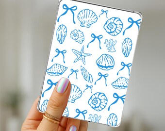 May include: A white phone case featuring a blue seashell and bow pattern. The design includes various seashell illustrations, starfish, and bows. The case is held in a hand with iridescent nail polish and gold rings.