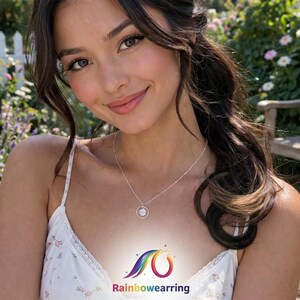 May include: A woman wearing a white floral print camisole and a silver necklace with a circular pendant. The image includes the text "Rainbowwearing" with a rainbow logo. The background features a garden with flowers and a wooden bench.