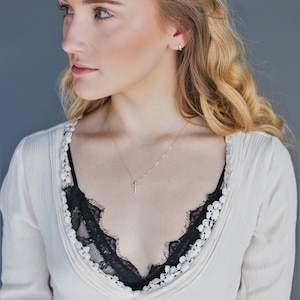 Könnte beinhalten: Eine Frau mit langen blonden Haaren trägt eine weiße Bluse mit schwarzer Spitzenborte und eine goldene Halskette mit einem kleinen Kreuzanhänger.