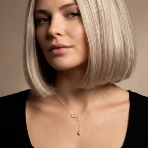 May include: A silver necklace featuring an infinity symbol and a heart-shaped pendant. The necklace is worn by a person in a black top. The background is a neutral beige colour.