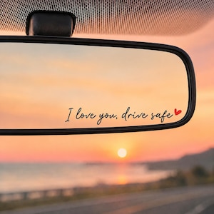 May include: A black car rearview mirror featuring a decal with the black cursive text I love you, drive safe and a small red heart icon. The mirror reflects a sunset over a road and water, with the sky showing orange and yellow hues.