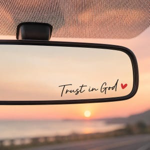 May include: A black vinyl decal applied to a car rearview mirror featuring the cursive black text Trust in God followed by a small red heart icon. The mirror reflects a sunset over a road and a body of water in the background.
