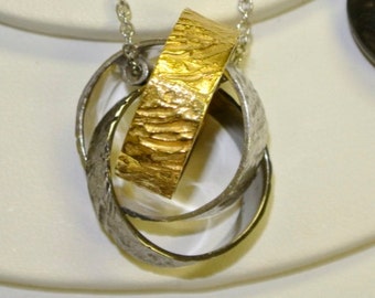 May include: A silver chain necklace with a pendant made of three interlocking rings. The center ring is gold and has a textured surface. The other two rings are silver.