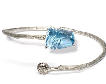 May include: A silver bangle bracelet with a light blue gemstone set in the center. The bracelet is designed to look like a branch with a textured surface.