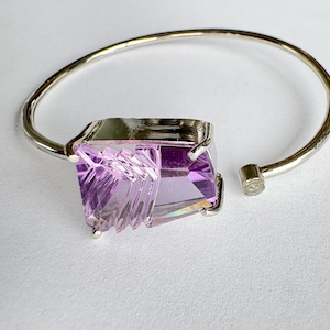 Könnte beinhalten: Offener Armreif in Silberoptik mit einem großen, facettierten, rechteckigen lila Edelstein. Der Edelstein ist in einer silbernen Lünette gefasst und hat einen vielseitigen Schliff. Das Armband ist an einem Ende offen mit einem kleinen, runden Silberverschluss.