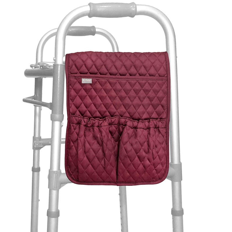 May include: A burgundy quilted walker bag with a diamond pattern. The bag has a top pocket and two lower pockets with gathered openings. It is attached to a silver walker frame.