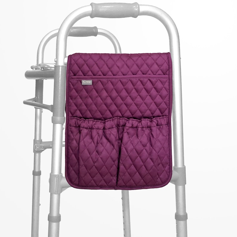 May include: A plum-coloured quilted walker bag with a diamond pattern. The bag features a zipped top pocket and two lower pockets with elastic openings. It is attached to a silver walker frame.