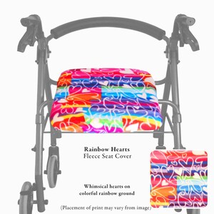 Puede incluir: Una funda de asiento de forro polar para andador con un fondo de rayas arco&iacute;ris vibrantes y un patr&oacute;n repetitivo de corazones blancos. El texto Rainbow Hearts Fleece Seat Cover aparece impreso debajo del asiento.