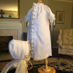 May include: White christening gown with lace trim and a matching bonnet. The gown is displayed on a wooden stand.