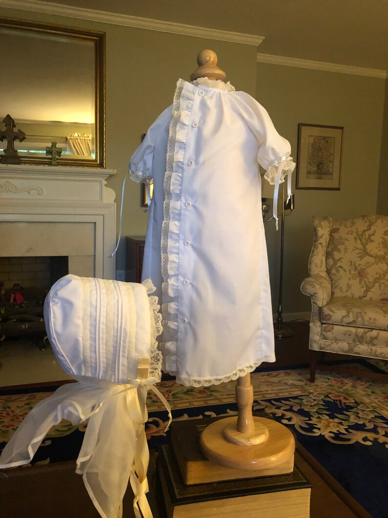 May include: White christening gown with lace trim and a matching bonnet. The gown is displayed on a wooden stand.