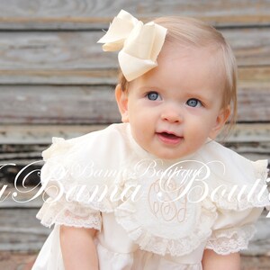 May include: A baby girl wearing a white dress with a lace trim and a large ivory bow in her hair. The dress has a white embroidered monogram on the front. The baby is looking up and smiling.