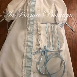 May include: A white baby gown with blue satin ribbon trim and lace. The gown has a ruffled collar and long sleeves with ties. A matching bonnet with blue satin ribbon trim and lace is included.