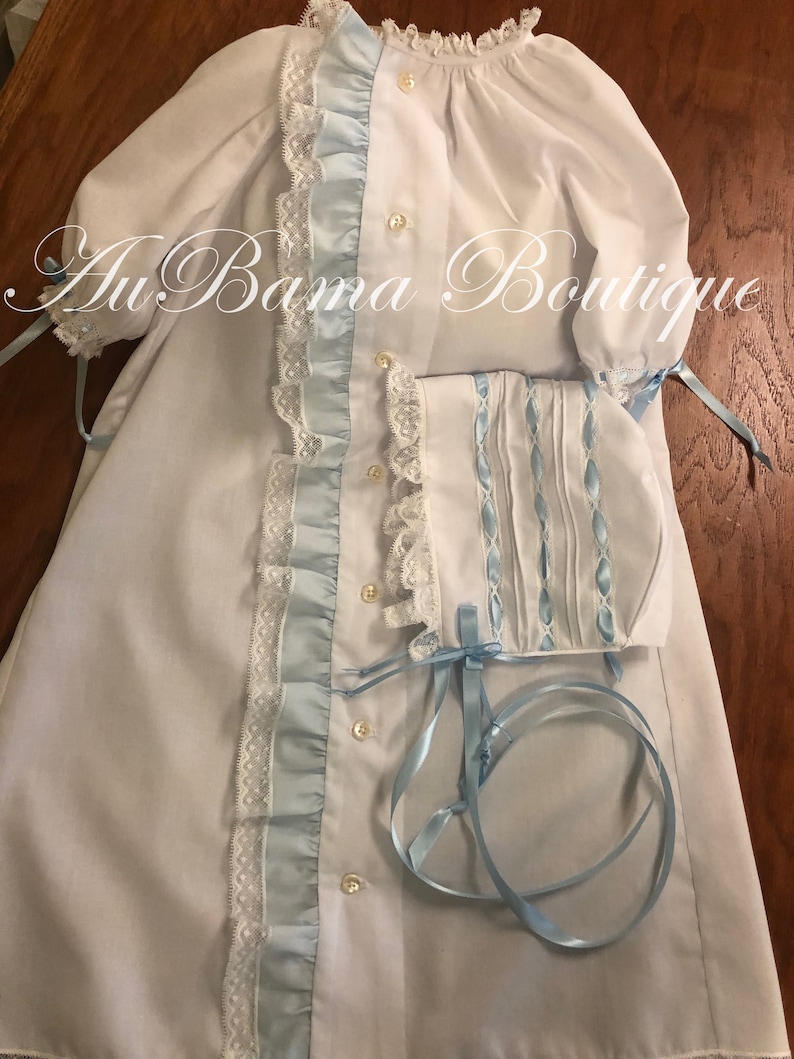 May include: A white baby gown with blue satin ribbon trim and lace. The gown has a ruffled collar and long sleeves with ties. A matching bonnet with blue satin ribbon trim and lace is included.