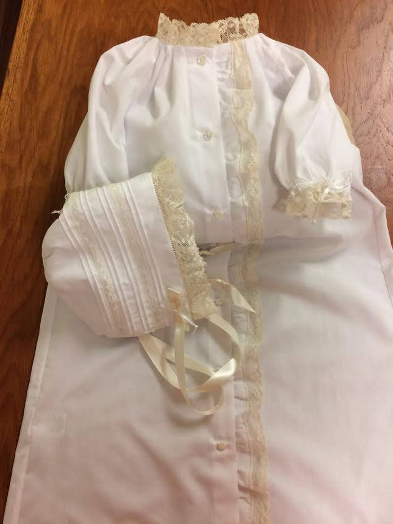May include: A white christening gown with a lace trim and a matching bonnet. The gown has a button-up front and long sleeves.