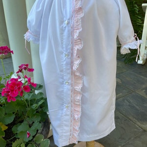 May include: A white baby gown with pink ruffled trim and a lace trim along the front. The gown has a button closure down the front.