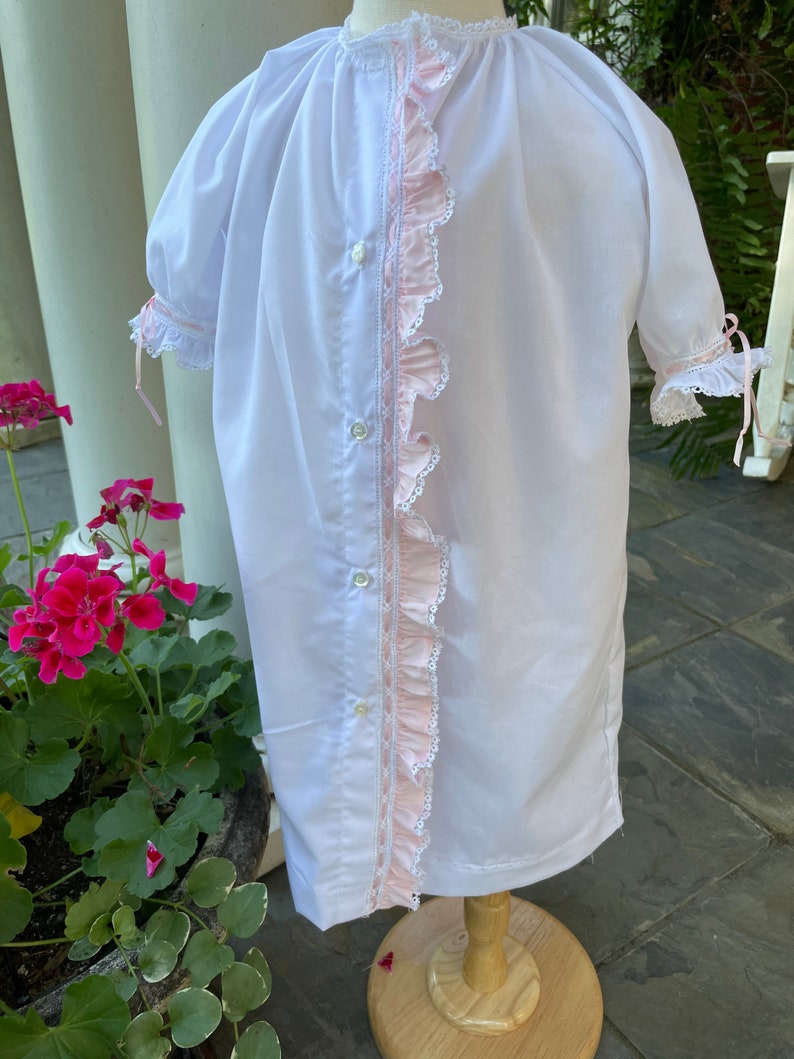 May include: A white baby gown with pink ruffled trim and a lace trim along the front. The gown has a button closure down the front.