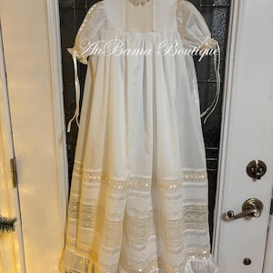 May include: White christening gown with lace trim and a ruffled hem. The gown has a high neck and short sleeves.