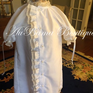 May include: A white christening gown with a lace trim and button closure. The gown is on a mannequin and has a ruffled trim down the side.