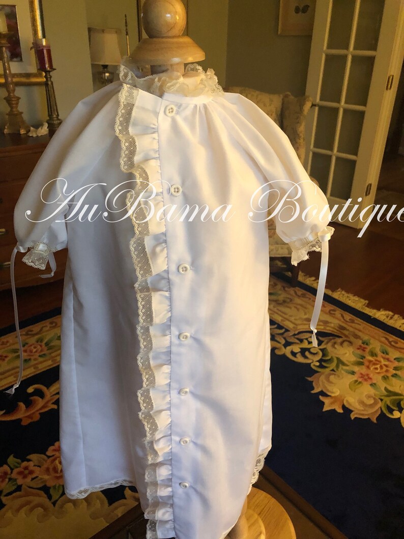 May include: A white christening gown with a lace trim and button closure. The gown is on a mannequin and has a ruffled trim down the side.