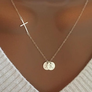 May include: A silver necklace featuring a sideways cross pendant and two small, round charms. The charms are engraved with the letters "J K". The necklace is displayed against a light-colored background.