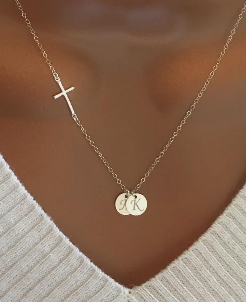 May include: A silver necklace featuring a sideways cross pendant and two small, round charms. The charms are engraved with the letters "J K". The necklace is displayed against a light-colored background.