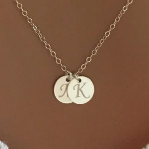 May include: A silver necklace with two circular pendants. Each pendant has a single initial engraved on it. The chain is a delicate link style, and the background is a solid brown.