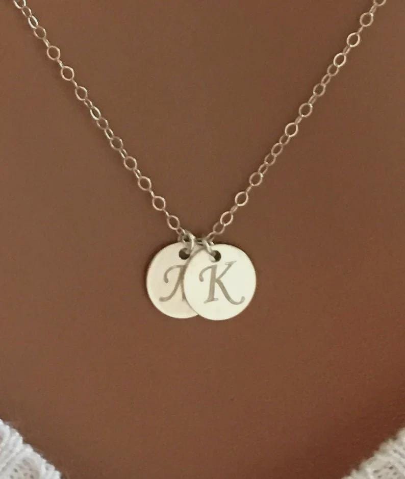 May include: A silver necklace with two circular pendants. Each pendant has a single initial engraved on it. The chain is a delicate link style, and the background is a solid brown.