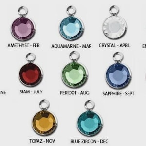 May include: A collection of twelve round, gemstone charms in various colors, each with a silver-colored loop at the top. Each charm is labeled with a month and gemstone name, including Siam, Amethyst, Aquamarine, Crystal, and Emerald.