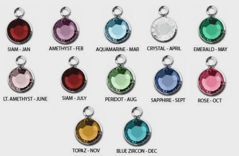 May include: A collection of twelve round, gemstone charms in various colors, each with a silver-colored loop at the top. Each charm is labeled with a month and gemstone name, including Siam, Amethyst, Aquamarine, Crystal, and Emerald.