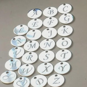 May include: A collection of silver-colored, circular charms, each engraved with a different letter of the alphabet. The charms are arranged in a grid pattern, with a small hole at the top of each for stringing. Some charms feature symbols.