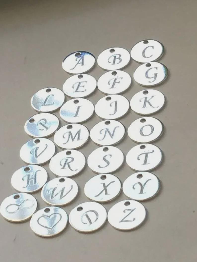 May include: A collection of silver-colored, circular charms, each engraved with a different letter of the alphabet. The charms are arranged in a grid pattern, with a small hole at the top of each for stringing. Some charms feature symbols.