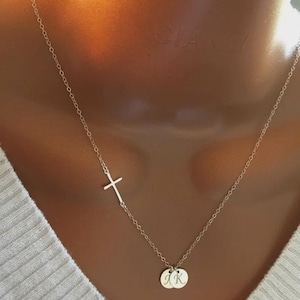 May include: A delicate silver necklace featuring a sideways cross pendant and two small, round charms. The charms appear to have initials engraved on them. The necklace is displayed against a neutral background.