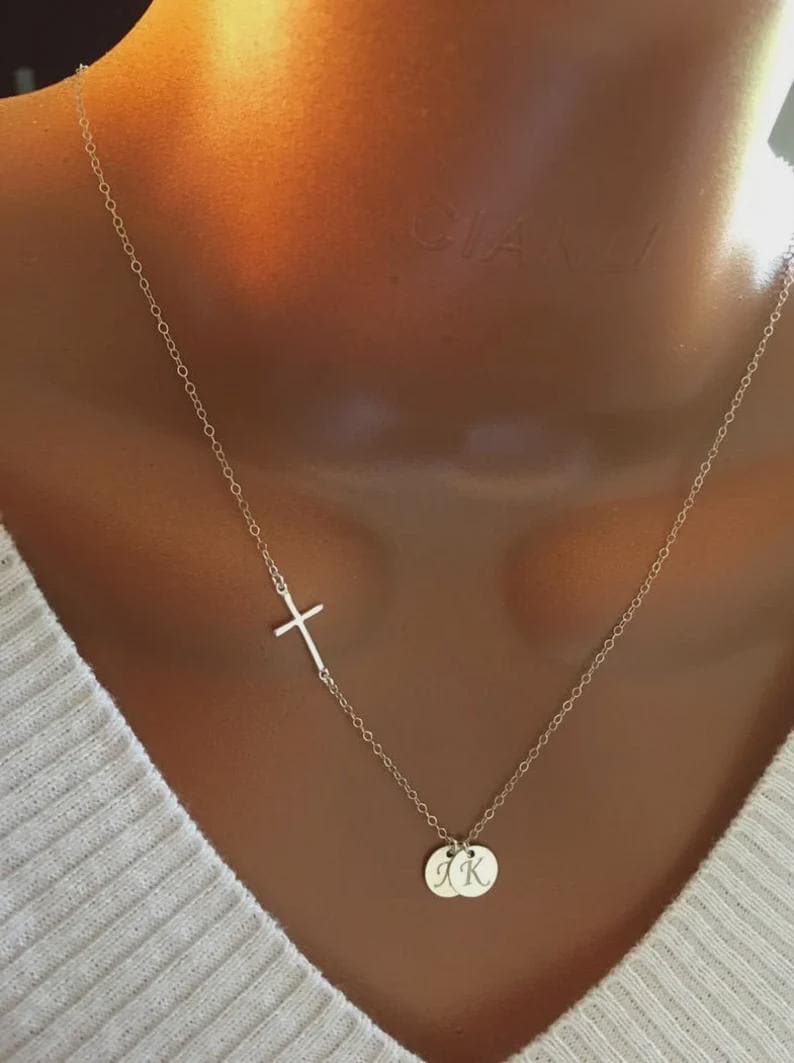 May include: A delicate silver necklace featuring a sideways cross pendant and two small, round charms. The charms appear to have initials engraved on them. The necklace is displayed against a neutral background.
