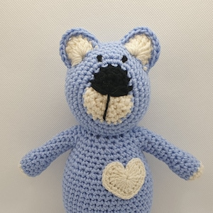Puede incluir: Un oso de peluche azul tejido a crochet con un corazón blanco en el pecho. El oso tiene una cara blanca con ojos negros y una nariz negra.