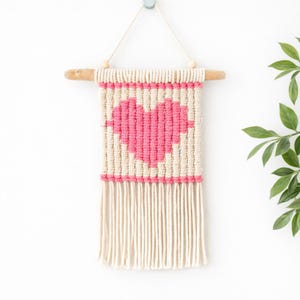 Könnte beinhalten: Makramee-Wandbehang mit einem rosa Herzdesign auf cremefarbenem Hintergrund. Das Herz befindet sich in der Mitte des rechteckigen Stücks, das unten einen Fransen hat. Aufgehängt an einer Holzstange mit Schnur und Holzperlen.