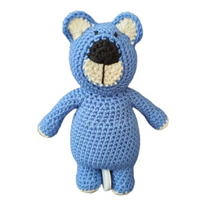 Puede incluir: Un oso azul tejido a mano con ganchillo, con nariz y boca negras, y detalles crema en las orejas, hocico y pies. El oso tiene una expresión amigable y brazos extendidos, ideal para un juguete infantil o decoración de guardería.