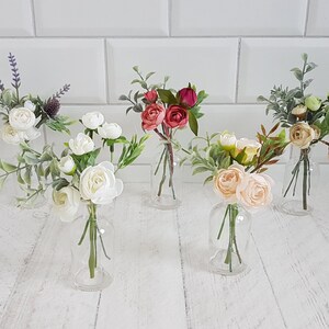 May include: Five small glass vases filled with artificial flower arrangements. The vases are clear glass and the flowers are white, pink, and red.