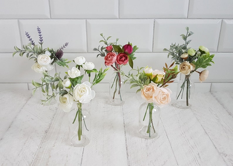 May include: Five small glass vases filled with artificial flower arrangements. The vases are clear glass and the flowers are white, pink, and red.