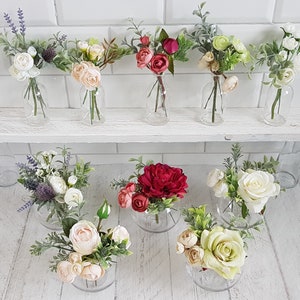 May include: Assortment of small floral arrangements in glass vases. The arrangements feature various flowers, including white, pink, and red roses, as well as greenery. The vases are clear glass and have a variety of shapes and sizes.
