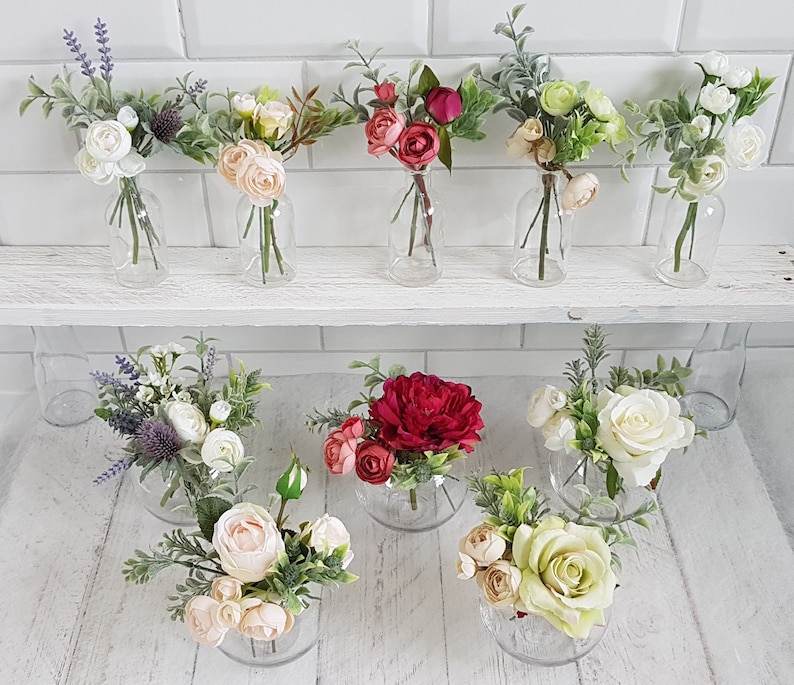 May include: Assortment of small floral arrangements in glass vases. The arrangements feature various flowers, including white, pink, and red roses, as well as greenery. The vases are clear glass and have a variety of shapes and sizes.